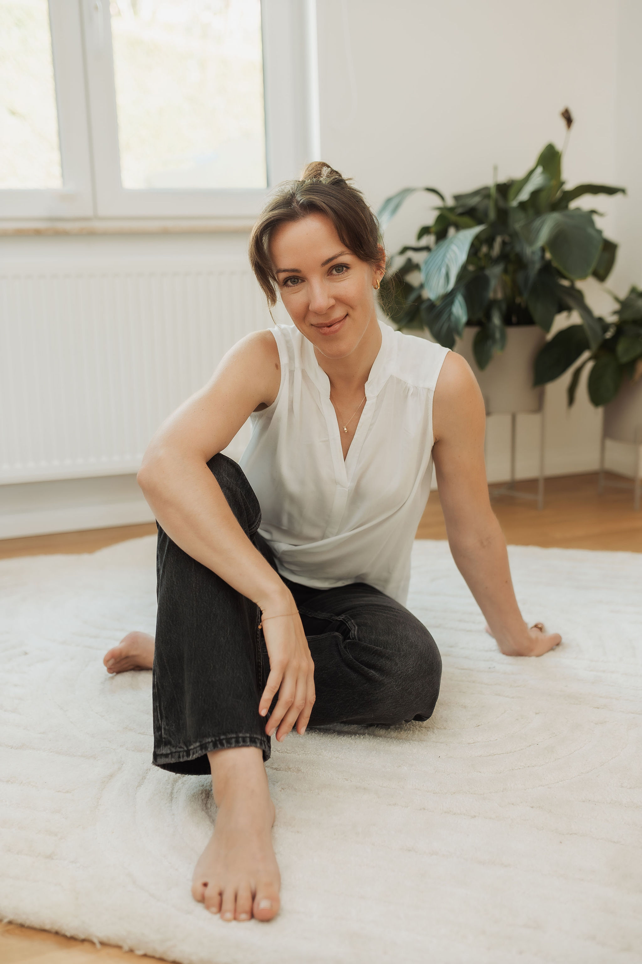 Eine Frau sitzt auf einem weißen Teppich in einem Raum mit einem Fenster im Hintergrund, trägt eine weiße Bluse und schwarze Hose, hat braune Haare und lächelt.