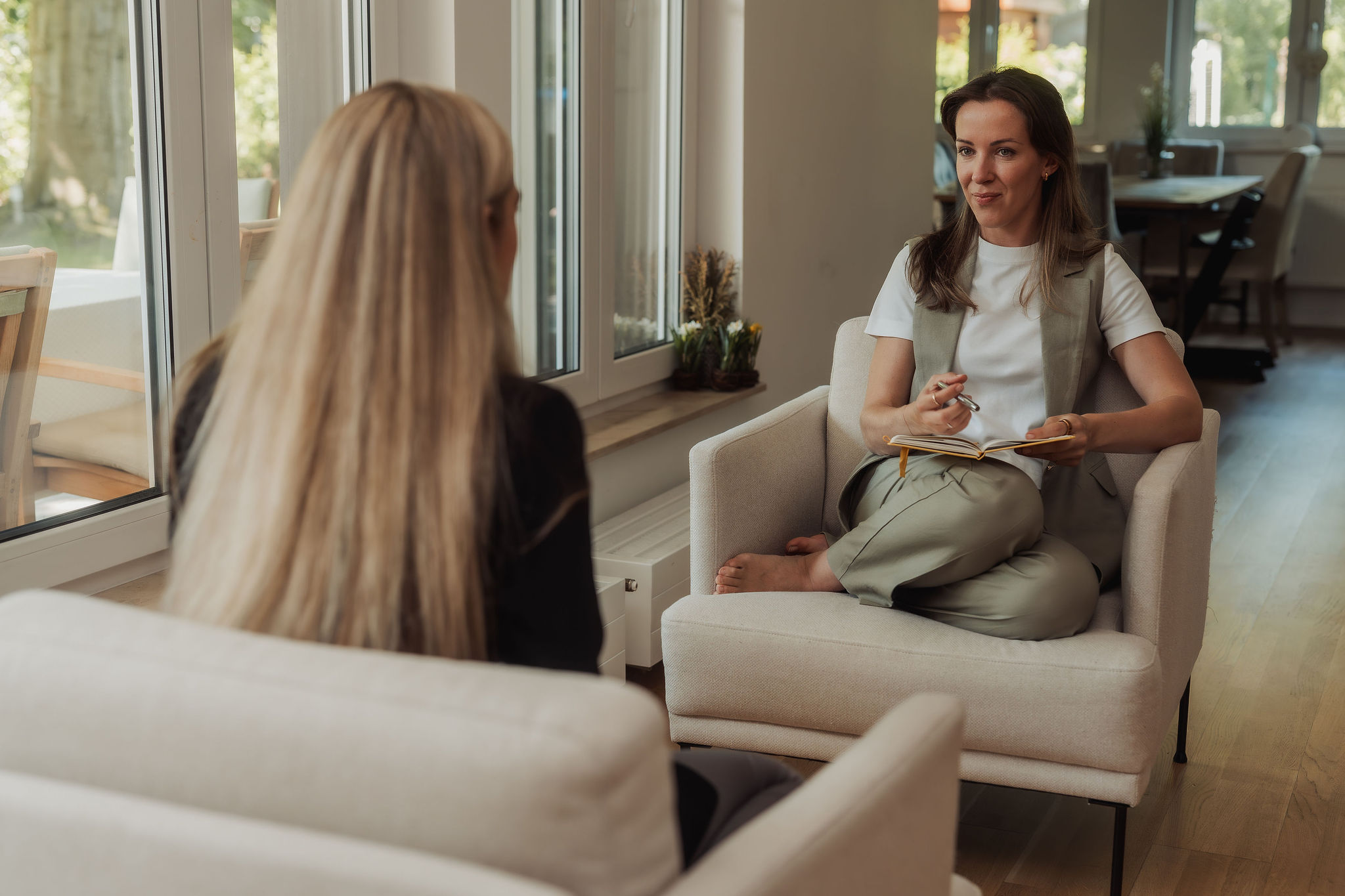 Zwei Frauen führen ein Gespräch in einem Wohnzimmer mit großen Fenstern und natürlichem Licht.
